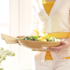 A woman carries a wooden salad bowl and tongs.
